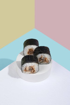 Téléchargez les photos : Hosomaki (sushis, rouleaux) avec anguille sur un support en plâtre blanc sur un fond uni coloré (bleu, rose, jaune). Une composition simple et concise - en image libre de droit