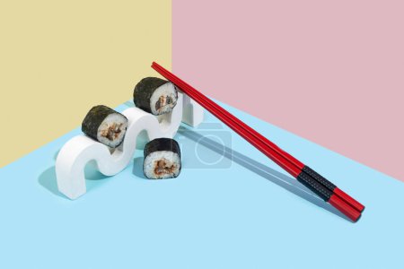 Photo for Hosomaki (sushi, rolls) with eel on a white plaster stand on a colorful plain background (blue, pink, yellow). A simple concise composition - Royalty Free Image