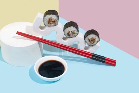 Téléchargez les photos : Hosomaki (sushis, petits pains) à l'anguille et sauce soja sur un support en plâtre blanc sur un fond uni coloré (bleu, rose, jaune). Une composition simple et concise - en image libre de droit