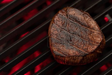 Téléchargez les photos : Un steak juteux grésille sur une grille en fonte au-dessus d'une flamme. Le steak est parfaitement cuit et a des marques de gril. Le fond est sombre et fumé - en image libre de droit