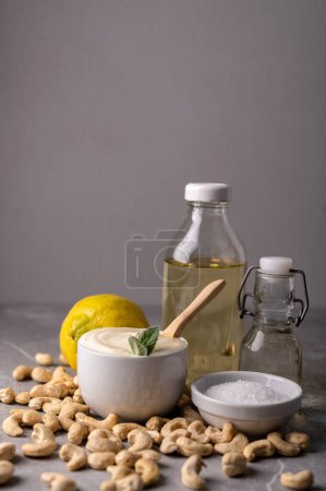 Photo for Cashew nut cream cheese in a white bowl and cooking ingredients - butter, salt, lemon in a plate on a gray marble stone background. This image represents a healthy snack option and can be used to convey concepts like nutrition, veganism, natural food - Royalty Free Image