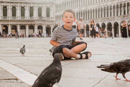 Photo for Cute seven-year-old boy in a striped t-shirt sits in St. Mark's Square in Venice and plays with pigeons. Tourism and recreation in Europe with children - Royalty Free Image