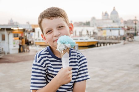 Téléchargez les photos : Mignon garçon de sept ans dans un t-shirt rayé se trouve sur le front de mer à Venise le soir et mange gelato (crème glacée). Tourisme et loisirs en Europe avec enfants - en image libre de droit