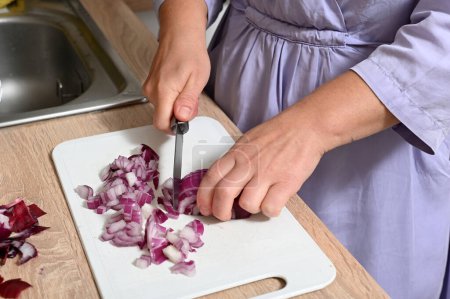 Téléchargez les photos : A woman cuts vegetables into squares. A woman peels red onions in her kitchen. Female hands hold a salad onion - en image libre de droit