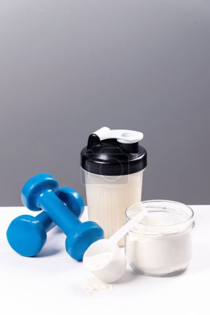 Sports nutrition: drink with proteins (collagen). A sports mixer, a glass and a dumbbell for fitness are on a white table