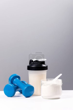 Sports nutrition: drink with proteins (collagen). A sports mixer, a glass and a dumbbell for fitness are on a white table