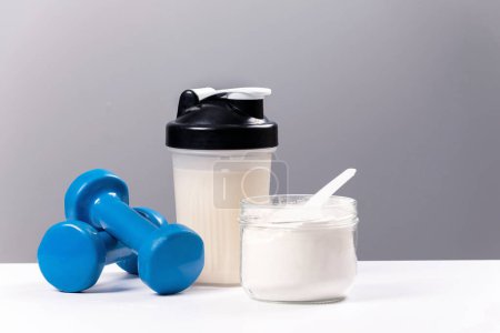 Photo for Sports nutrition: drink with proteins (collagen). A sports mixer, a glass and a dumbbell for fitness are on a white table - Royalty Free Image