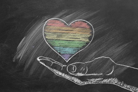 Foto de Una mano dibuja un corazón colorido con rayas de tiza arco iris sobre un fondo de pizarra oscura, que representa el amor y la diversidad. LGBT, concepto de orgullo - Imagen libre de derechos