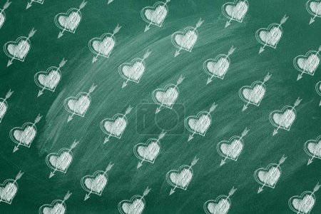 Foto de Una pizarra de aula verde muestra un patrón caprichoso de corazones atravesados por flechas, todos dibujados en tiza, creando una escena lúdica y romántica. - Imagen libre de derechos