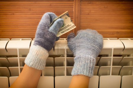 Foto de Las manos en manoplas de punto cálido se calientan en el radiador con dinero en las manos. - Imagen libre de derechos