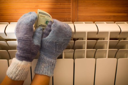 Foto de Las manos en manoplas de punto cálido se calientan en el radiador con dinero en las manos. - Imagen libre de derechos