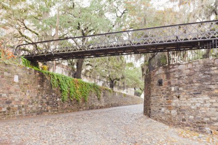 Téléchargez les photos : Cobblestone road, stone walls and iron pedestrian foot-bridge of River Street Access in Historic District of downtown city of Savannah, Georgia, GA, USA - en image libre de droit
