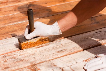 Photo for Painter staining deck boards with transparent protective outdoor decking paint applying stain with brush with special attention to the gaps - Royalty Free Image