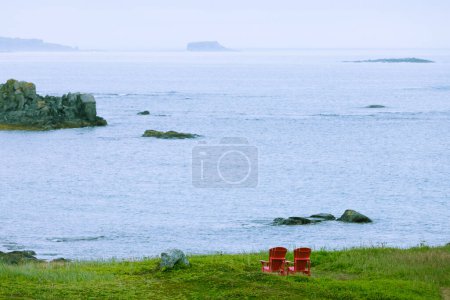 Photo for Red wooden Adirondack Chairs in beautiful coastal landscape of Strait of Belle Isle, Newfoundland, NL, Canada - Royalty Free Image