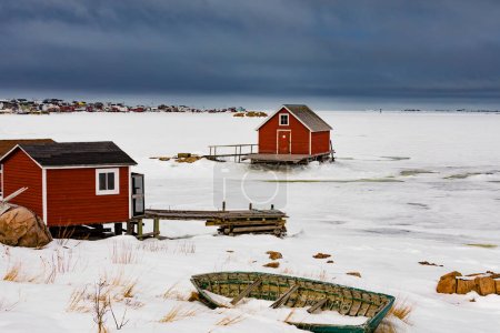 Fishing stage shacks and old wooden skiff row boat at shore of frozen North Atlantic Ocean in outport town of Joe Batt's Arm on Fogo Island, Newfoundland, NL, Canada