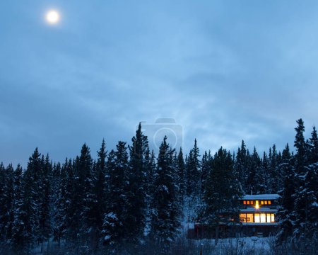 Cozy country living home warmly illuminated isolated in boreal forest taiga moon-lit frozen winter landscape of remote Yukon Territory, YT, Canada