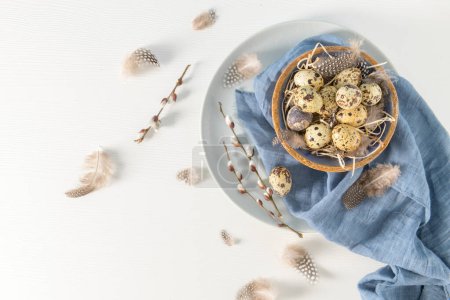 Quail eggs in a ceramic bowl with a blue scarf and bird feathers. Wooden table painted white. Background with elements suggestive of the Easter season.