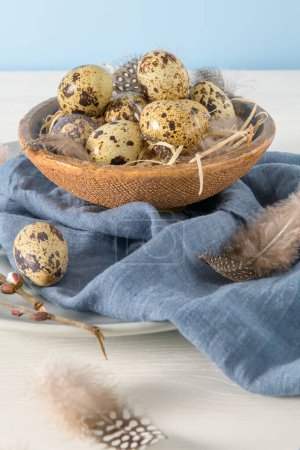 Quail eggs in a ceramic bowl with a blue scarf and bird feathers. Wooden table painted white and a plain blue background. Background with elements suggestive of the Easter season.