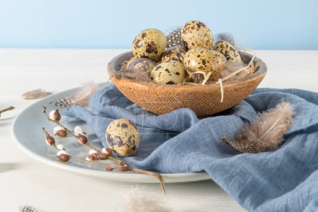 Quail eggs in a ceramic bowl with a blue scarf and bird feathers. Wooden table painted white and a plain blue background. Background with elements suggestive of the Easter season.