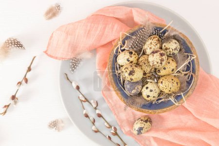 Quail eggs in a ceramic bowl with a peach fuzz pink colored scarf and bird feathers. Wooden table painted white. Background with elements suggestive of the Easter season.
