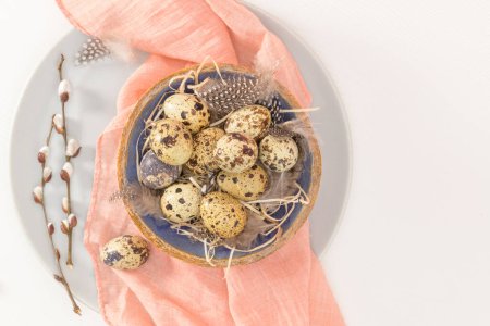 Quail eggs in a ceramic bowl with a peach fuzz pink colored scarf and bird feathers. Wooden table painted white. Background with elements suggestive of the Easter season.