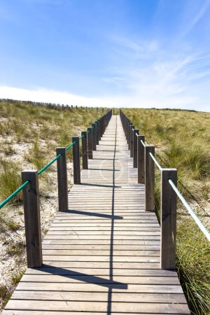 A wooden boardwalk through sand dunes leads to a coastal town of Vila Praia de Ancora nestled against hills