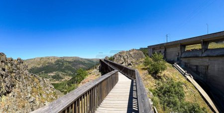 A wooden walkway leading to a dam, surrounded by rugged terrain under a clear blue sky. Guarda, Portugal.