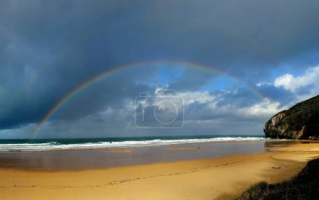 A stunning seascape with a vibrant rainbow arching over a tranquil beach, kissed by gentle waves. The dramatic sky adds depth to the scene, creating a mesmerizing and serene view.