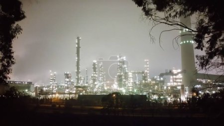 Foto de Vista nocturna de un complejo industrial que brilla con luces, contrastando con un árbol solitario en primer plano, mezclando elementos urbanos y naturales en una escena cautivadora. - Imagen libre de derechos