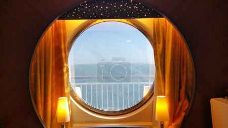Experience luxury aboard a cruise ship in this elegantly designed cabin with a large round window, golden curtains, a sofa, two armchairs, and a coffee table. Perfect for a relaxing vacation getaway.