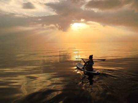 Eine junge Frau im Bikini paddelt bei Sonnenuntergang auf einem ruhigen See. Die Farben des Himmels spiegeln sich im Wasser wider, während sie die friedliche Szenerie genießt und sich der Natur verbunden fühlt.