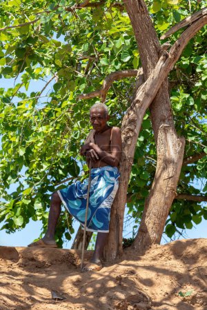 Foto de BELLO SUR TSIRIBIHINA, MADAGASCAR - 04.11.2022: Anciano cansado descansa a la sombra cerca de un ferry que cruza el río en Belo Sur Tsiribihina, Madagascar. - Imagen libre de derechos
