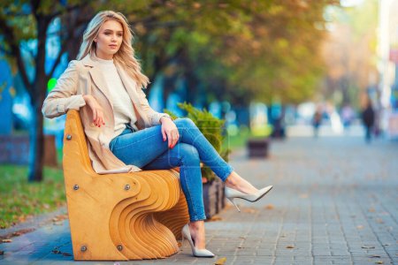 Horizontales Porträt einer schönen jungen kaukasischen Blondine, die entspannt auf einer Holzbank sitzt und blaue Jeans und beigen Trenchcoat mit unscharfem blaugelben Herbsthintergrund trägt.