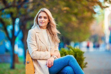 Nahes Porträt einer lächelnden schönen jungen kaukasischen Blondine, die entspannt auf einer Holzbank sitzt und blaue Jeans und beigen Trenchcoat mit unscharfem bläulich-gelbem Hintergrund trägt