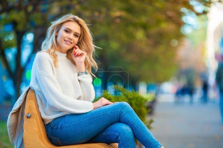 Nahes Porträt einer lächelnden schönen jungen kaukasischen Blondine, die entspannt auf einer Holzbank sitzt und blaue Jeans und weißen Pullover mit verschwommenem blaugelben Hintergrund der Herbststadt trägt.