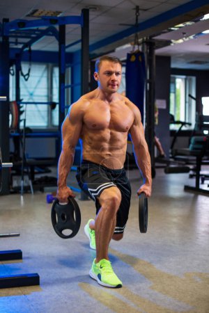 Athletic fit sporty muscular caucasian man holding two weight disc plates in his hands while doing lunges topless in black shorts in contemporary gym, blurred bluish background