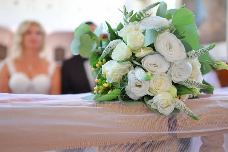 Photo for Wedding bouquet of white flowers on the railing of the kneeler in the church during the wedding. Young couple in the background. - Royalty Free Image