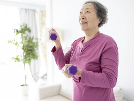 Foto de Asiática senior mujer haciendo ejercicios con mancuernas en el interior. - Imagen libre de derechos
