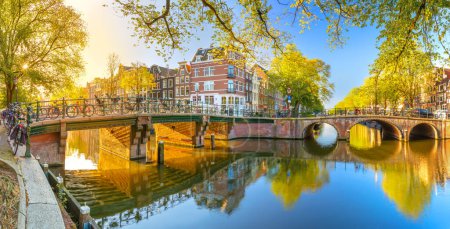 Panoramic view of Amsterdam in the morning sun. Traditional old houses, bridges and mirror water with reflection. Beautiful autumn morning in Amsterdam. Holland, Netherlands, Europe.