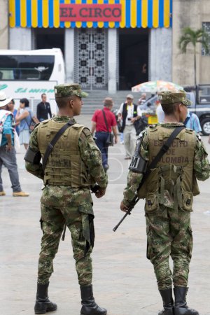 Foto de MEDELLIN, COLOMBIA - 14 DE MARZO: Dos militares de pie en la plaza con el arma automática en las manos y las personas alrededor. El Circo último cartel de exhibición en el fondo. Colombia 2015 - Imagen libre de derechos
