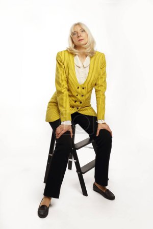 Belle dame blonde dans la cinquantaine portant une veste jaune posant sur fond blanc