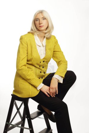Beautiful blond lady in her 50s wearing yellow jacket posing over white background