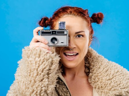 Téléchargez les photos : Belle rousse tenant un appareil photo 35mm vintage avec flash cube sur fond bleu - en image libre de droit