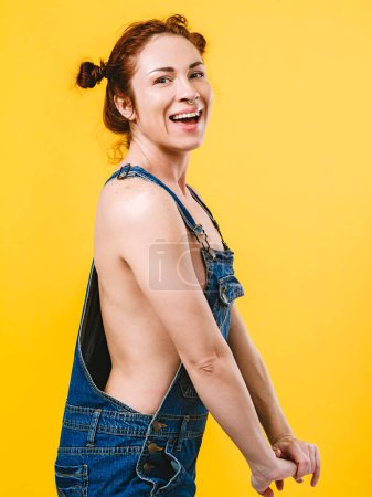 Téléchargez les photos : Sexy rousse seins nus femme riant et portant des salopettes sur fond jaune. - en image libre de droit