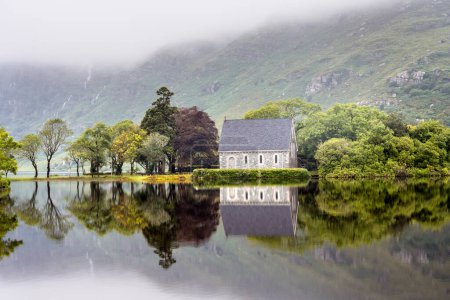 Kleine Kirche, die sich in ruhigem Wasser spiegelt. St. Finbarrs Oratorium befindet sich auf einer Insel im See in Gougane Barra, Irland.