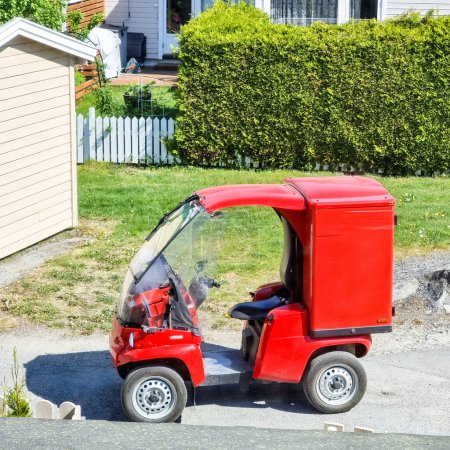 Tiny red car with a red container, parked in front of a garden in a small street of neighbourhood a sunny summer day.