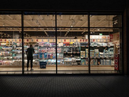 Téléchargez les photos : Un regard à travers une fenêtre dans un supermarché la nuit. Un homme se tient debout avec un téléphone portable. Copenhague, Danemark - 14 août 2023. - en image libre de droit