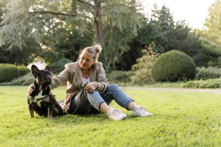 Photo for Blonde girl of forty years old cuddles with her dog, a French bulldog, in a park on the grass. - Royalty Free Image