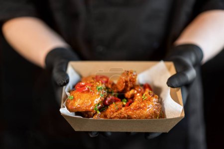 Photo for The cook holds in his hands a box with grilled spicy chicken teriyaki legs in packs for delivery. - Royalty Free Image
