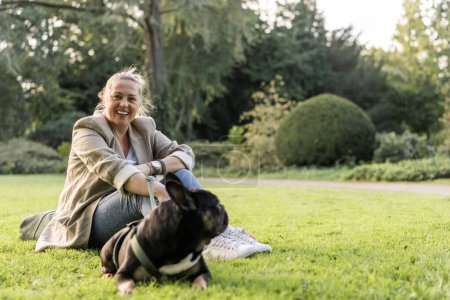Téléchargez les photos : Une blonde de quarante ans se blottit avec son chien, un bouledogue français, dans un parc sur l'herbe. - en image libre de droit